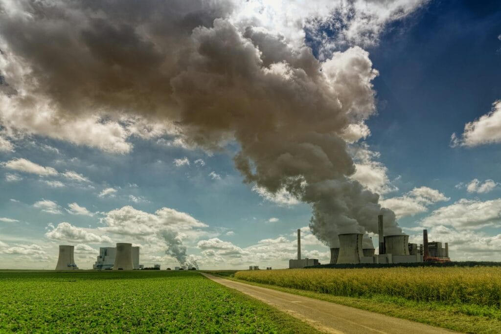 Power plant in rural landscape emitting smoke under a bright blue sky with fluffy clouds.