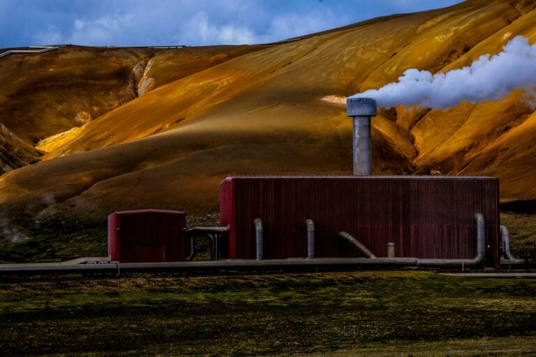 Red geothermal power plant emitting steam against a dramatic yellow-brown mountainous backdrop. Steam Power Plants
